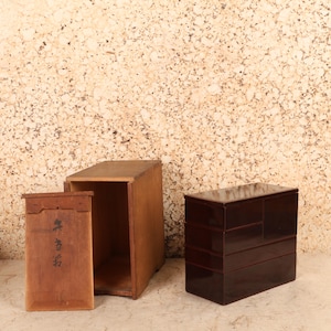 May include: A rectangular wooden storage box with a detached lid featuring black ink calligraphy, positioned next to a dark brown, multi-tiered lacquerware chest with a smooth, glossy finish and several stacked drawers of varying sizes.