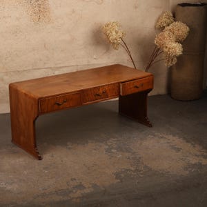 May include: A wooden desk featuring a smooth, rectangular top with rounded edges and three drawers across the front. Each drawer is equipped with a curved, dark metal handle. The desk is supported by two solid, continuous side panels.