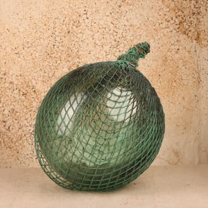 May include: A spherical glass fishing float encased in a green knotted rope net. The glass is translucent with a pale green tint, and the netting features a gathered knot at the top, creating a textured, protective covering over the round surface.
