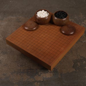 May include: A wooden Go board featuring a grid of thin black lines, accompanied by two matching wooden bowls filled with white and black stones respectively. Two circular wooden lids are placed on the board surface next to the bowls.
