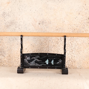 Puede incluir: Un soporte de exhibición de madera negra con un diseño decorativo, que sostiene un objeto de madera marrón claro. El soporte presenta una escena de paisaje con incrustaciones de nácar. El soporte está diseñado para sostener un objeto largo y cilíndrico.