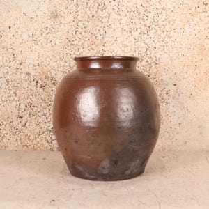 May include: A large, brown ceramic jar with a rounded body and a slightly flared rim. The jar has a smooth, glossy finish with variations in color, ranging from light to dark brown. The jar is likely used for storage or decorative purposes.