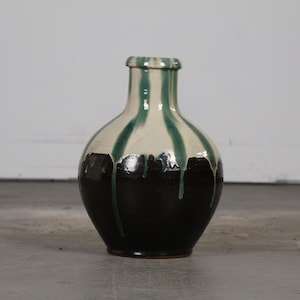 May include: A ceramic vase with a rounded body and a narrow neck. The vase is primarily black at the base, transitioning to a cream color with green vertical stripes. The green paint drips down the sides, creating a unique design. The vase is likely for decorative purposes.