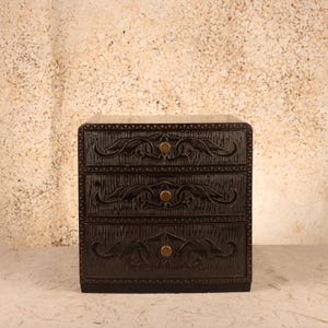 May include: A dark brown wooden chest with three drawers, each featuring a carved leaf-like pattern and a single circular brass knob. The front surface of the chest has a textured, vertical grooved finish, and the edges are detailed with a small, repeating notched border design.