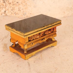 Può includere: Un piccolo tavolo in legno con un piano di colore marrone scuro e dettagli dorati. Il tavolo presenta intagli intricati sui lati e sulle gambe, ed è decorato con foglia d'oro. Il tavolo è perfetto per esporre piccoli oggetti o come pezzo decorativo.