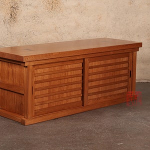 May include: A wooden cabinet with two sliding doors. The doors have a slatted design and are made of light brown wood. The cabinet is sitting on a concrete floor.