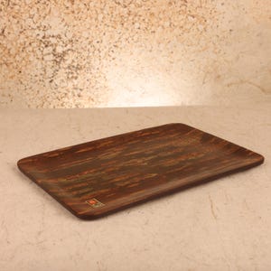 May include: A rectangular tray with a dark brown wood grain finish and a smooth surface. The tray features a small, square, multi-colored logo sticker in the bottom left corner, showcasing a distinct pattern of dark brown and lighter brown streaks.