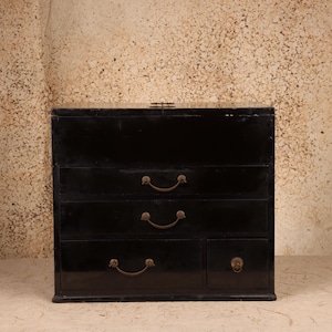 May include: A black lacquered wooden chest featuring a rectangular top compartment with a metal latch, two full-width drawers, and a lower section containing one wide drawer and one smaller square drawer, all fitted with curved metal pull handles.