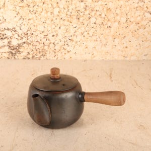 May include: A dark metallic teapot with a rounded body, a short spout, and a wooden side handle. The lid features a small wooden knob at the center. The surface of the teapot shows a smooth, matte finish with subtle signs of wear and dark patina.