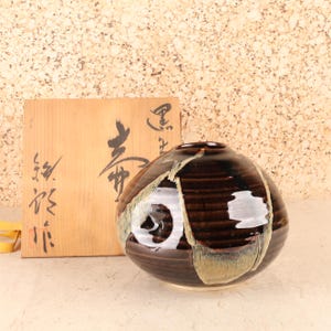 Könnte beinhalten: Eine runde, dunkelbraune Keramikvase mit glänzender Oberfläche und hellen, strukturierten Glasurakzenten. Neben der Vase steht eine quadratische Aufbewahrungsbox aus Holz, die mit schwarzen japanischen Kalligrafiezeichen beschriftet ist.