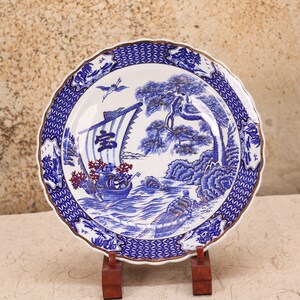 May include: A decorative ceramic plate with a blue and white Japanese design. The plate features a boat, trees, and a bird. The rim has a scalloped edge with a gold trim. The plate is displayed on a wooden stand.