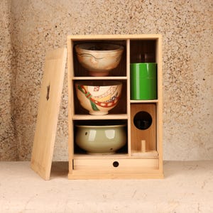 May include: A light-colored wooden tea cabinet with a removable door, featuring three stacked ceramic bowls with varied patterns, a bright green cylindrical tea canister, a small wooden stand with a circular cutout, and a bottom storage drawer.