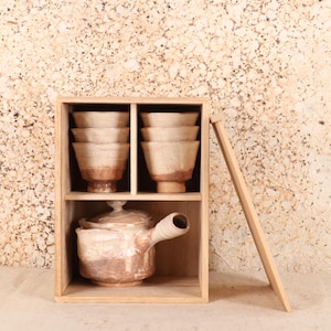 Puede incluir: Una caja de madera con dos estantes, que contiene seis cuencos de cerámica y una tetera. Los cuencos están apilados en pares, y la tetera tiene un asa y una tapa. La cerámica es de color marrón claro y crema.