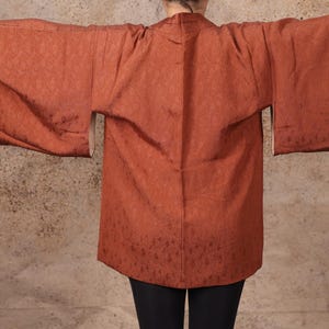 May include: A rust-colored kimono-style jacket with wide sleeves. The jacket has a subtle floral pattern and a contrasting white lining visible at the sleeve openings. The garment is worn over black pants.