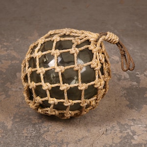 May include: A spherical, dark green glass float encased in a hand-knotted, light tan jute rope net. The netting features a square grid pattern and includes a sturdy, braided rope loop extending from the top for hanging or attachment purposes.