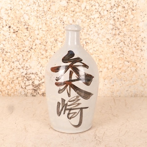 Puede incluir: Una botella de sake de cerámica de color crema con un cuerpo redondeado y un cuello estrecho. La botella presenta caligrafía japonesa en tinta marrón y negra. El fondo es una superficie texturizada de color claro.