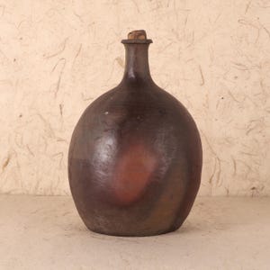 Puede incluir: Una botella de color marrón oscuro, de cuerpo redondo, con cuello largo y tapón de corcho. La botella tiene un acabado liso y brillante y un tono marrón rojizo en el centro. La botella es probablemente de cerámica o gres.