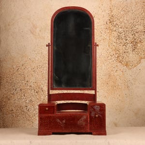 May include: A red vanity mirror with a tall, arched glass pane mounted on a swivel frame. The base features a small drawer on the left, a larger central drawer, and a cabinet on the right, all decorated with etched crane and floral motifs.