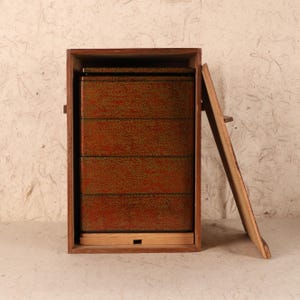 May include: A wooden, multi-tiered bento box with a reddish-brown finish and green speckles. The box is housed in a larger wooden frame, with the lid slightly ajar. The design suggests a traditional Japanese aesthetic, ideal for serving or storing food.