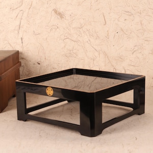 May include: A black lacquered square table with a glass top. The table has a gold emblem on the side. The table is a low, rectangular design, suitable for a living room or a similar setting.