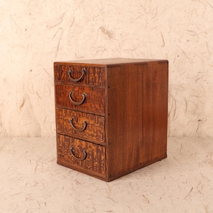 May include: A small, wooden chest of drawers with four drawers. The wood has a warm, reddish-brown tone with a unique grain pattern. Each drawer has a curved, metal handle. The chest is a compact size, suitable for small spaces.