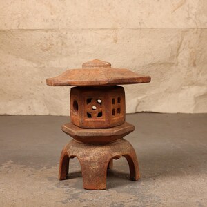 May include: A terracotta-colored stone lantern featuring a hexagonal central chamber with intricate cut-out geometric patterns, a wide overhanging roof, and a sturdy base supported by four arched legs, showcasing a weathered and textured surface.