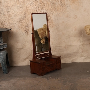 May include: A wooden vanity mirror with a rectangular frame that pivots on two side supports. The base features a central drawer and two side compartments, all crafted from dark wood with visible grain patterns and small decorative cutouts.