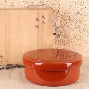 Puede incluir: Un recipiente redondo lacado en rojo con tapa, frente a una caja de madera clara. La caja tiene un asa tejida y caracteres japoneses. El recipiente es probablemente para té o almacenamiento de alimentos.