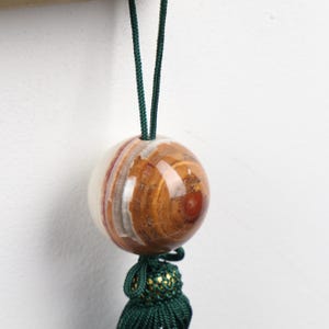 May include: A decorative, spherical ornament with a polished, banded stone appearance. The sphere is suspended by a dark green cord and features a matching tassel with gold accents. The stone has a mix of white, brown, and reddish-brown hues.