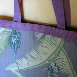 Puede incluir: Primer plano de un bolso tote con un estampado floral. El bolso presenta un fondo morado claro con grandes hojas verdes y blancas. La parte superior y las asas son de color morado liso. El bolso es de tela.