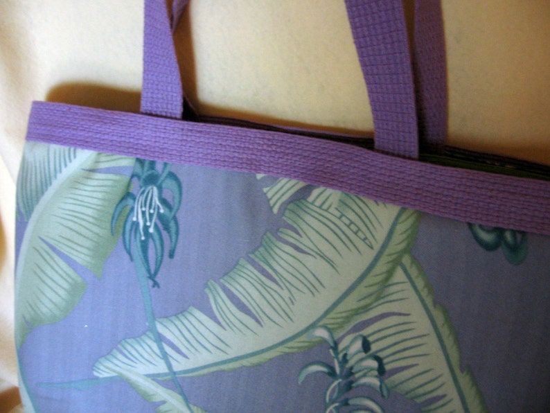 Puede incluir: Primer plano de un bolso tote con un estampado floral. El bolso presenta un fondo morado claro con grandes hojas verdes y blancas. La parte superior y las asas son de color morado liso. El bolso es de tela.