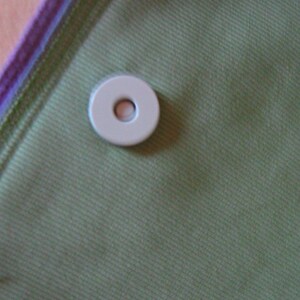 Puede incluir: Primer plano de un bot&oacute;n blanco circular con un agujero en el centro, sobre una tela verde. La tela tiene una sutil textura de costillas diagonales. El borde de la tela est&aacute; ribeteado con morado y verde.
