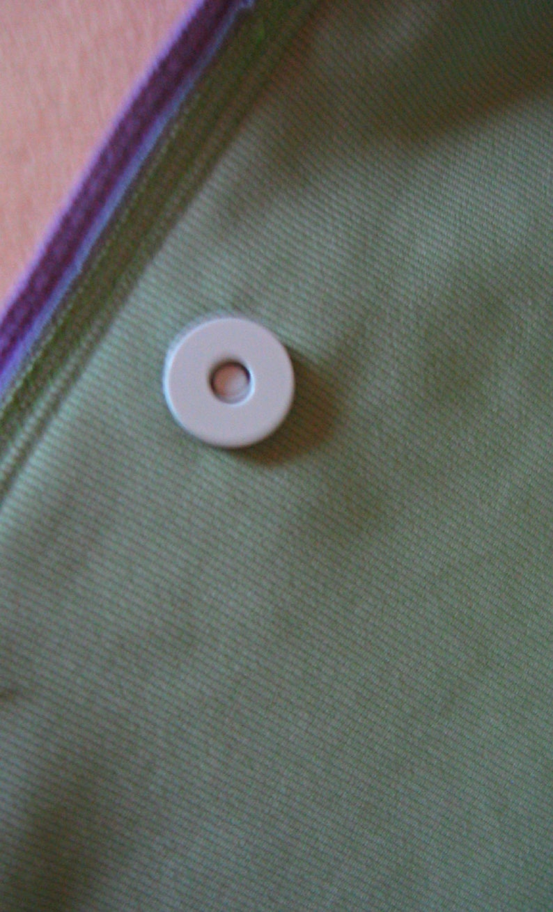 Puede incluir: Primer plano de un bot&oacute;n blanco circular con un agujero en el centro, sobre una tela verde. La tela tiene una sutil textura de costillas diagonales. El borde de la tela est&aacute; ribeteado con morado y verde.
