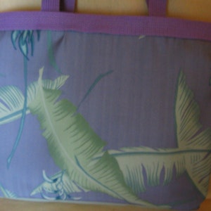 Puede incluir: Un bolso tote con un fondo lavanda y un estampado de hojas tropicales en tonos verdes y azules. El bolso tiene un ribete y asas lavanda.