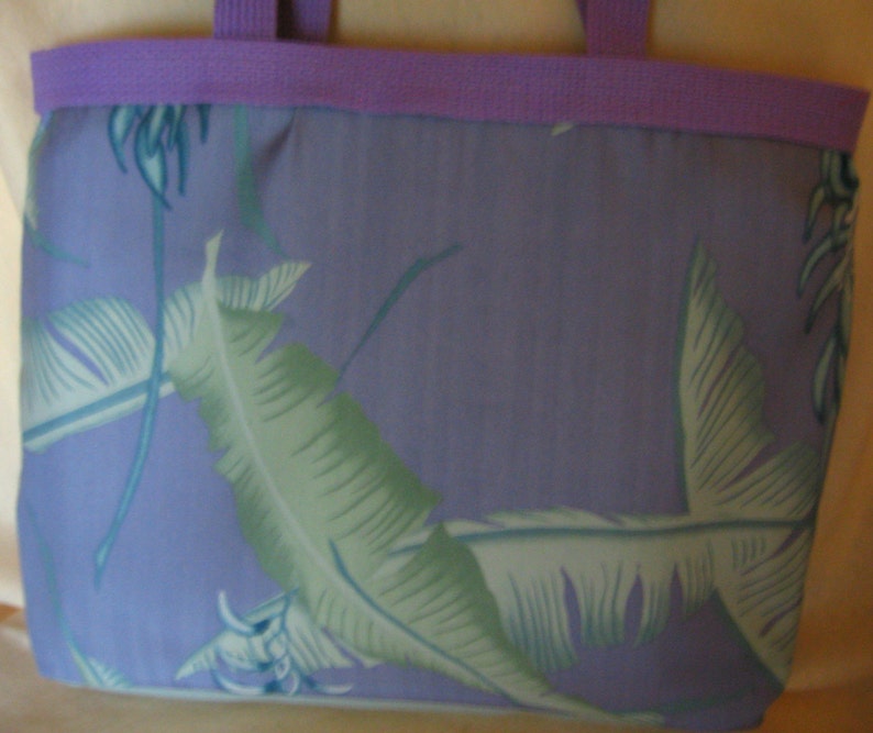 Puede incluir: Un bolso tote con un fondo lavanda y un estampado de hojas tropicales en tonos verdes y azules. El bolso tiene un ribete y asas lavanda.