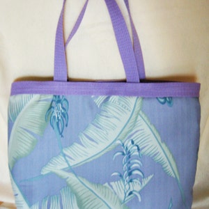 Puede incluir: Un bolso tote con ribete y asas lilas. El bolso presenta un estampado de hojas tropicales azules y blancas sobre un fondo azul claro. Las asas son de un material tejido resistente.