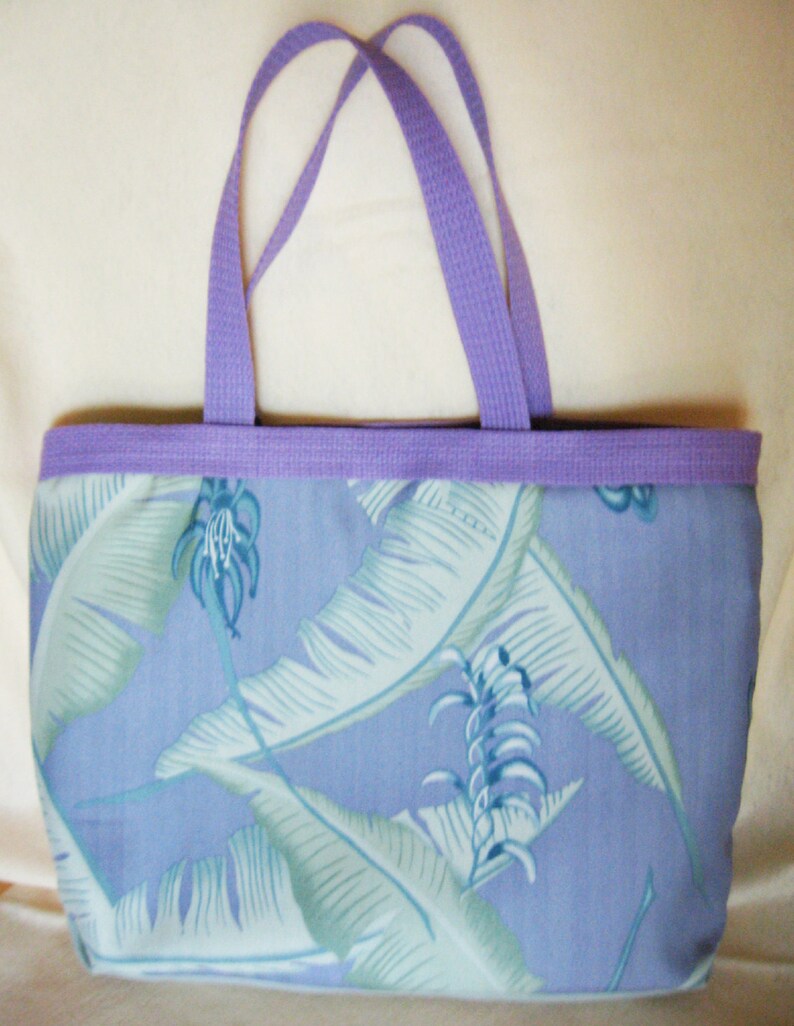Puede incluir: Un bolso tote con ribete y asas lilas. El bolso presenta un estampado de hojas tropicales azules y blancas sobre un fondo azul claro. Las asas son de un material tejido resistente.