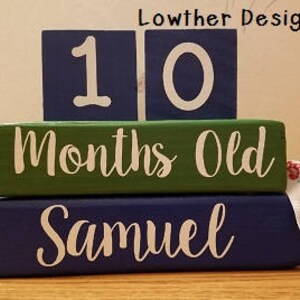 May include: Wooden blocks for milestone photos. The top blocks read "10" in white on a navy blue background. The middle block is green and says "Months Old" in white. The bottom block says "Samuel" in white on navy blue.