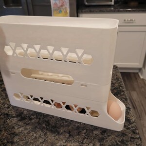 May include: A white plastic egg holder, rectangular in shape, with geometric cutouts and a dispensing tray. Several eggs are visible inside the holder and one egg is in the dispensing tray.