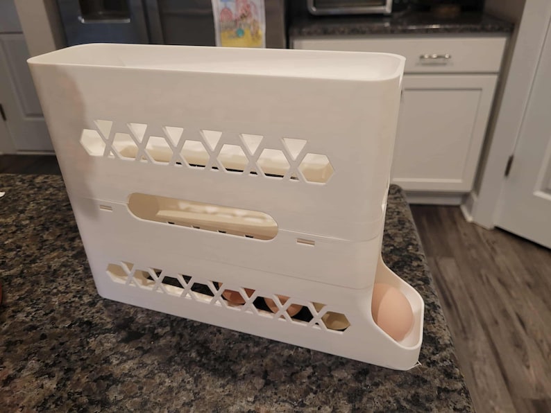 May include: A white plastic egg holder, rectangular in shape, with geometric cutouts and a dispensing tray. Several eggs are visible inside the holder and one egg is in the dispensing tray.