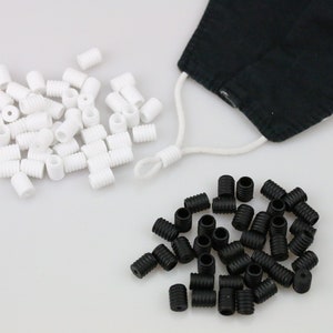 May include: An image showing white and black cord locks, used for adjusting face masks. A black face mask with white cords is also visible. The cord locks are on a white surface.