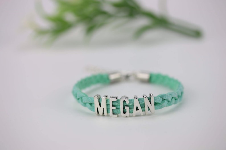 May include: A braided turquoise leather bracelet with a silver charm that spells out the name "MEGAN".