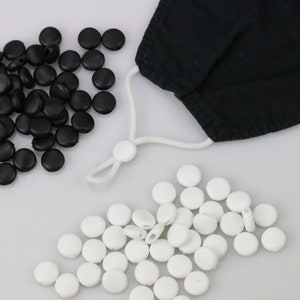 May include: A close-up of black and white plastic mask strap adjusters. The adjusters are round and have a small hole in the centre. They are arranged in two piles on a white surface.