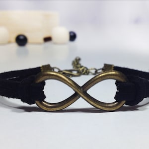 May include: A black suede bracelet with an infinity symbol charm. The charm is made of bronze and has a slightly aged finish.