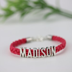 May include: A red braided cord bracelet with a silver charm that spells out the name "MADISON".