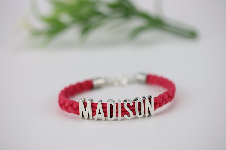 May include: A red braided cord bracelet with a silver charm that spells out the name "MADISON".