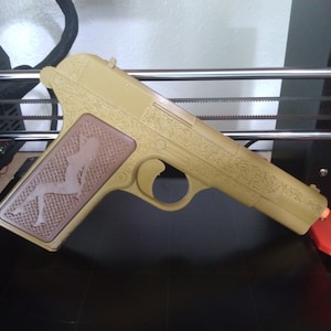 May include: A 3D printed, tan-colored plastic gun with a detailed floral design. The grip is brown and features a silhouette of a woman.