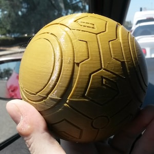 Puede incluir: Una esfera amarilla con una superficie texturizada y un patrón geométrico. La esfera está siendo sostenida en la mano de una persona.