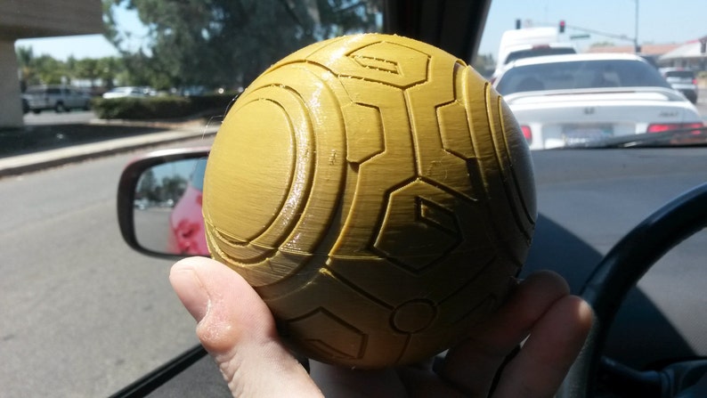 May include: A yellow sphere with a textured surface and a geometric pattern. The sphere is being held in a person's hand.