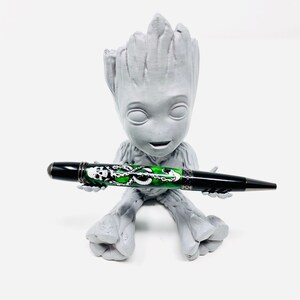 Può includere: Una figura grigia di Groot stampata in 3D che tiene una penna nera e verde. La penna presenta un design a teschio e tentacoli. La penna è nera con dettagli argentati. La figura di Groot è di colore grigio chiaro.